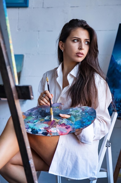 Painter paints with watercolors in workshop at home Portrait of pretty young woman artist in art studio and painting on canvas