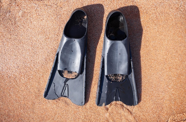 A pair of black flippers on the background of sand next to the water, top view. Swimming equipment