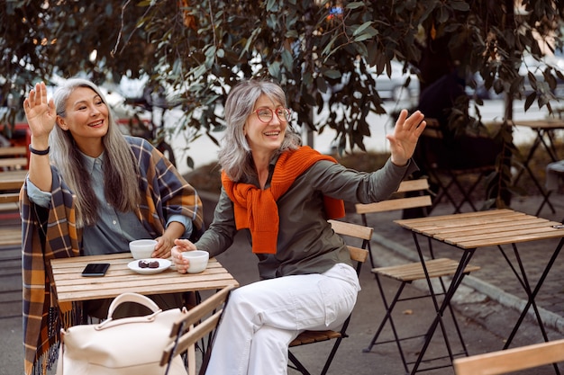 Pair of mature women friends wave hand calling waiter in street cafe