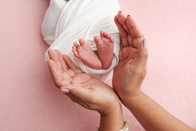 The palms of the parents father and mother hold the legs feet of a newborn baby in a white wrapper on a pink background Feet heels and toes of a newborn child close up Professional macro photo