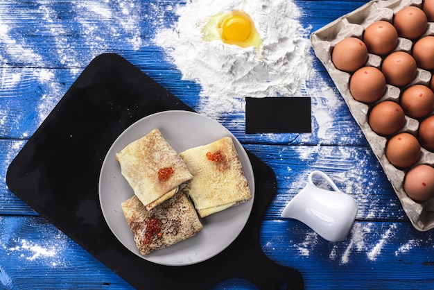 Pancakes on a blue wooden background with seafood on top of red caviar, flour is scattered next to it, a raw egg is broken and a plate with raw eggs