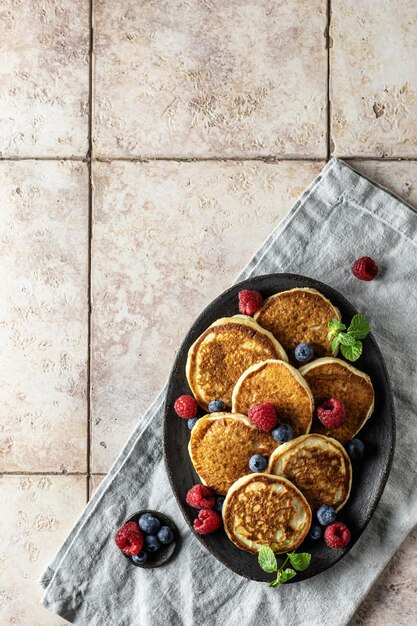 Pancakes in brown ceramic plate with raspberries blueberries mint leaves on linen napkin on a pink tile background top view