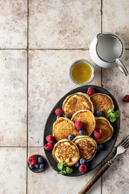 Pancakes in ceramic plate with raspberries blueberries mint leaves honey and fork on a pink tile background top view