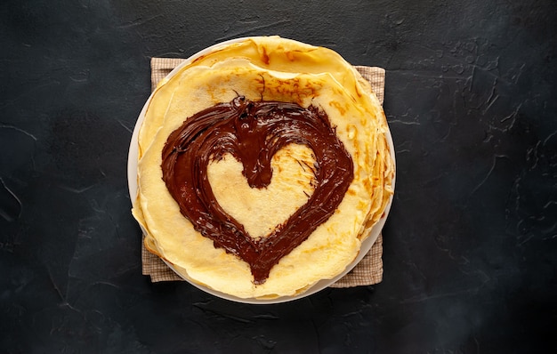 Pancakes with chocolate paste and hazelnuts, a heart is drawn on pancakes on a white plate on a stone background