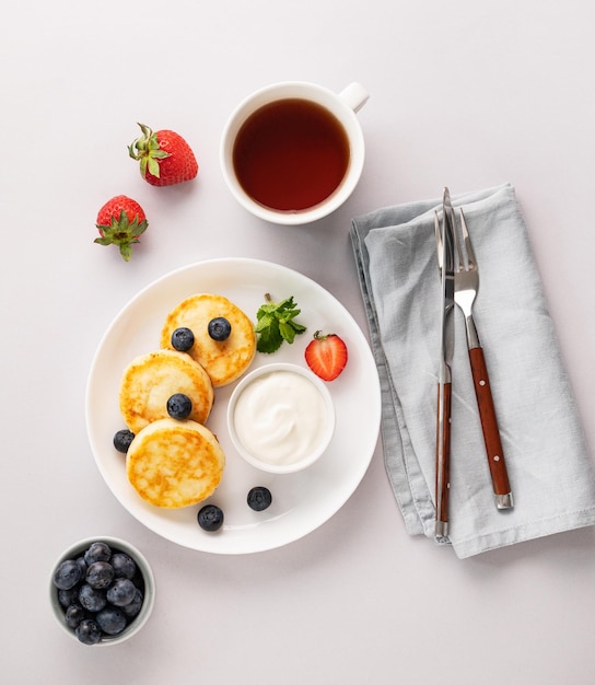 Pancakes with cottage cheese with fresh blerries and sour cream on a gray background with a cup of tea The concept of a healthy and delicious breakfast Top view