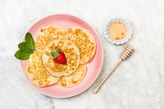 Pancakes with one strawberry on top on a pink plate and on a marble table in a top view