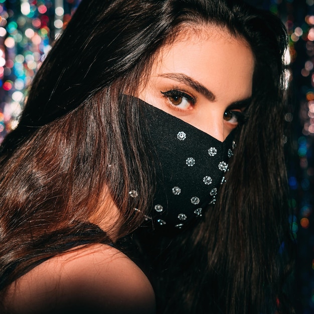 Pandemic beauty. Quarantine accessory. Party look. Portrait of mysterious brunette woman in black glamour creative design fabric face mask with crystal decor on blur shiny background.