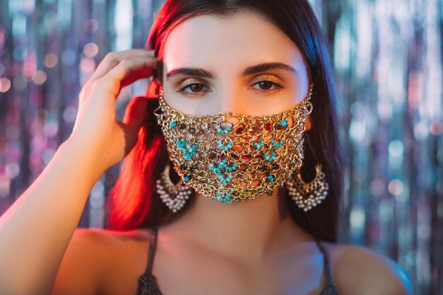 Pandemic fashion. Christmas party. Quarantine festive accessory. Portrait of tender woman wearing glamour gold chain face mask in red blue neon glow on bokeh light blur shiny tinsel background.