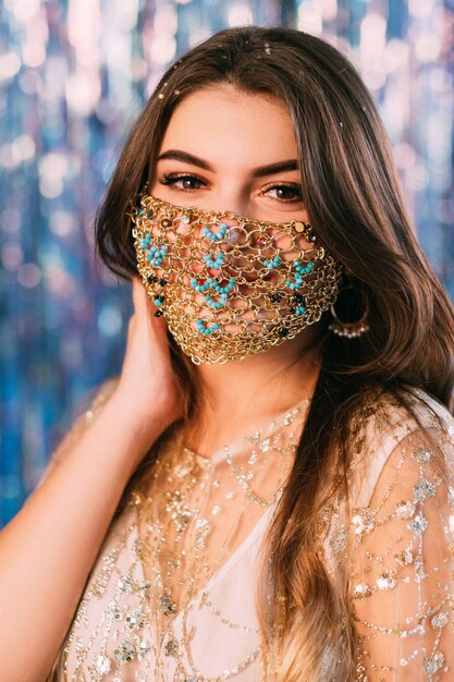 Pandemic fashion. Christmas party. Quarantine festive jewelry. Portrait of tender woman wearing glamour oriental style gold chain face mask on blue pink bokeh light blur shiny tinsel background.