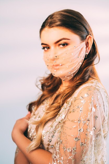 Pandemic fashion. Party look. Quarantine special occasion. Tender female model in sequin mesh face mask matching golden silver cocktail dress posing isolated on pastel pink light background.