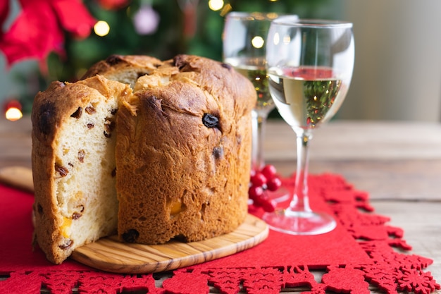 Panettone and white wine glasses on a wooden table with christmas decorations and tree in the backgr...