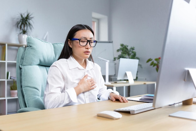 Panic attack in the workplace young asian woman sitting at desk in office holding chest breathing