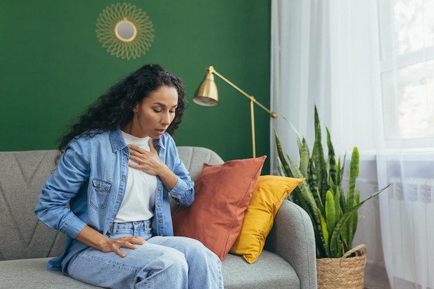 Panic attack a young beautiful latin american woman is sitting at home on the sofa holding her chest