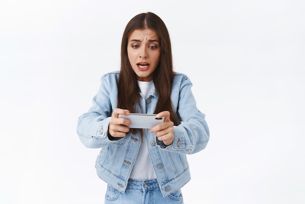 Panicking young intense cute brunette geeky girl in denim jacket playing fighting game or shooter using mobile phone holding smartphone horizontally grimacing as feeling worried want pass level