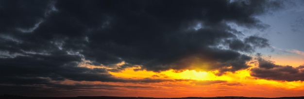 Panorama of dramatic colorful sunset with dark and bright clouds. The sun's rays are breaking through the cloudy sky. Dark clouds against a bright saturated sky.