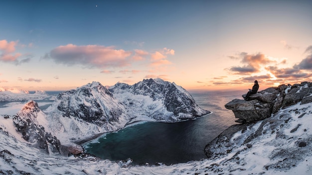 Panorama of Mountaineer sitting on rock on peak mountain of arctic coastline at sunset. Ryten Mountain, Lofoten islands, Norway