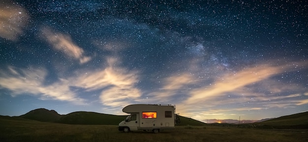 Panoramic night sky over Campo Imperatore highlands, Abruzzo, Italy. The Milky Way galaxy arc and stars over illuminated camper van. Camping freedom in unique hills landscape.