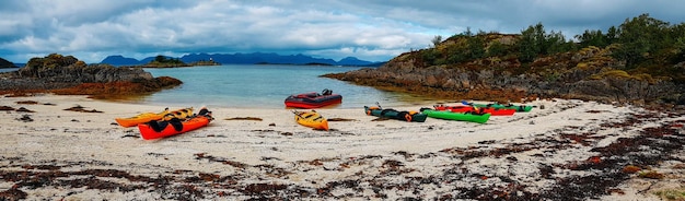 Panoramic photo of colorful kayaks on the sandy seashore hills blue sky in Norway