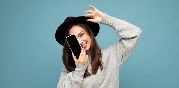 Panoramic photo of Pretty young smiling woman wearing black hat and grey sweater holding phone looking at camera isolated on background.Mock up, cutout, free space. copy space