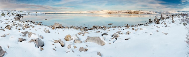 Panoramic view of norwegian lake at sunset. There are stone columns on rocks covered with snow.