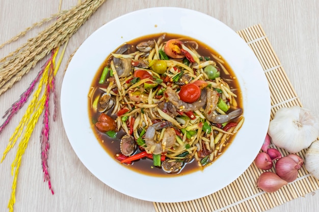 Papaya salad in a white plate is placed on the table and garlic rice, onions and ears of rice are placed around it.