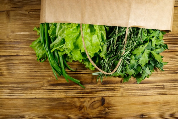 Paper bag with green onion rosemary lettuce leaves and parsley on wooden table Top view Healthy food and grocery shopping concept