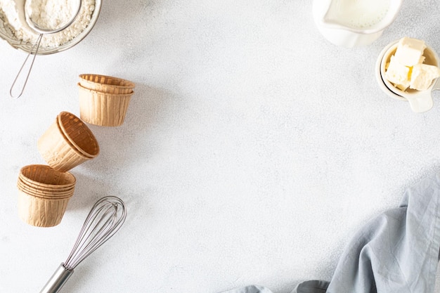 Paper cupcake baking pans on a white concrete background and ingredients Flour butter sugar whisk milk Copy space Top view Baking concept flat lay