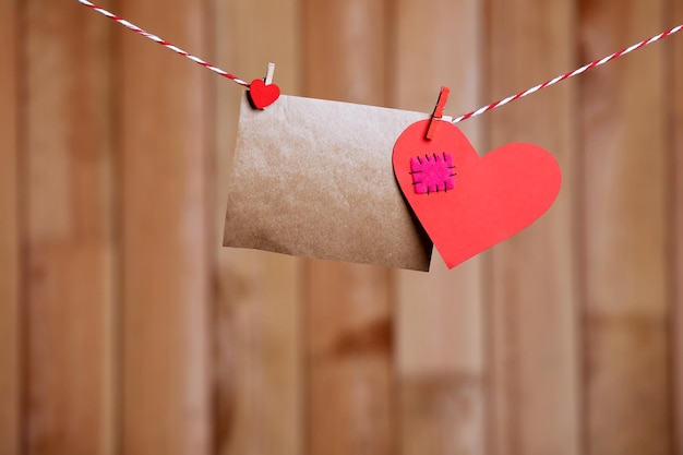 Paper hearts and empty sheet hanging on cord against wooden background