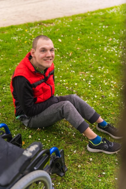 A paralyzed young man sitting on the grass next to the wheelchair next to a tree looking at the camera