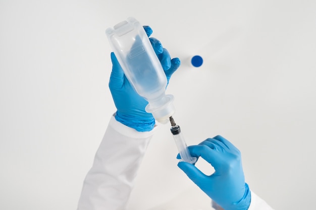 A paramedic on a white surface in blue latex gloves is taking a solution for separating powder medicine from a large transparent bottle with a syringe