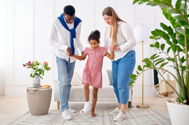 Parents help a little girl with a broken leg, living room interior. Mother, father and their little daughter cope with the trouble together, parental care