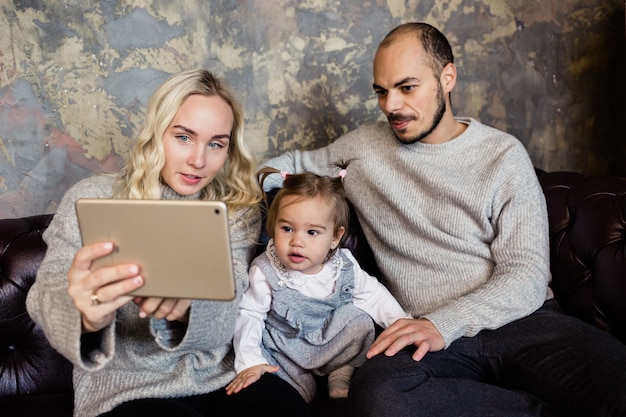 Parents with girl kid sitting on coach at home and talking with friends at video chat via tablet during Coronavirus disease