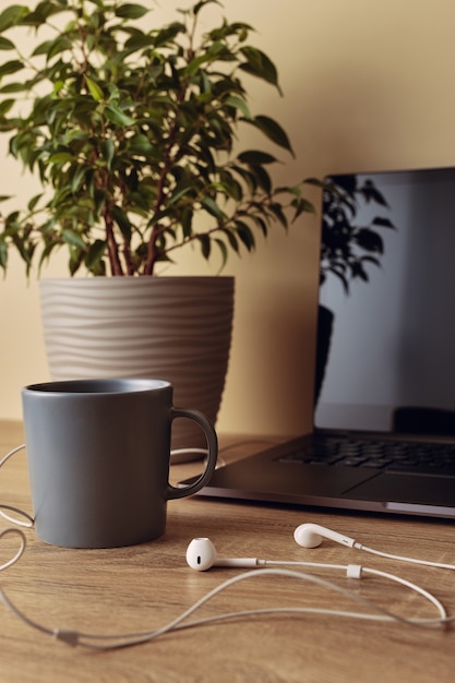 Part of laptop with blank screen, potted plant, mug and white cable headphones.