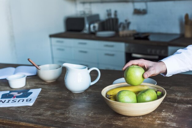 Partial view of man taking apple from bowl on wooden kitchen table