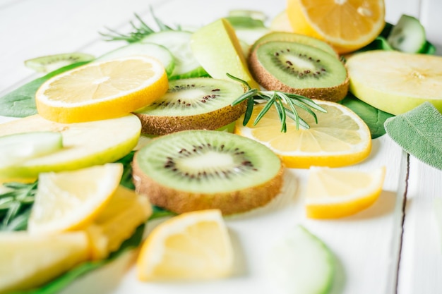 Partially blurred green and yellow fruits and herbs cut into pieces lie on white table Ingredients for smoothie fruit salad or dessert Slices of lemon apple kiwi cucumber spinach and tarragon