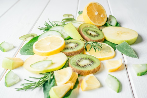 Partially blurred green and yellow fruits and herbs cut into pieces lie on white table Ingredients for smoothie fruit salad or dessert Slices of lemon apple kiwi cucumber spinach and tarragon