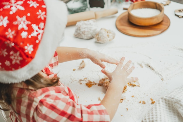 Partially blurred hands of little girl in Christmas cap and checkered shirt knit dough for cookies on white table