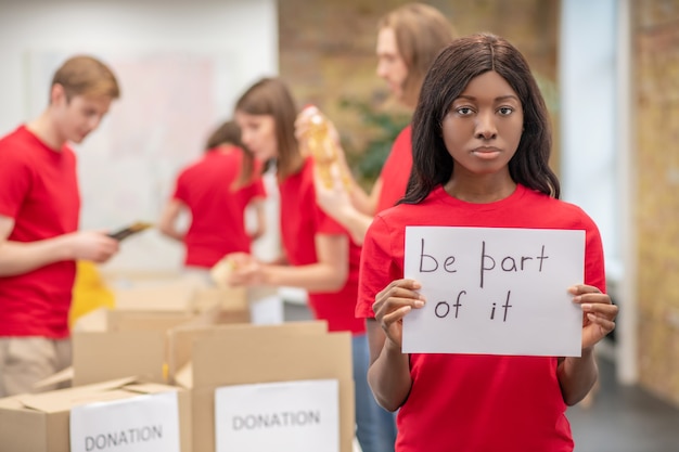 Participation, volunteering. Young focused american woman wearing red volunteer tshirt showing poster with slogan to help people in need