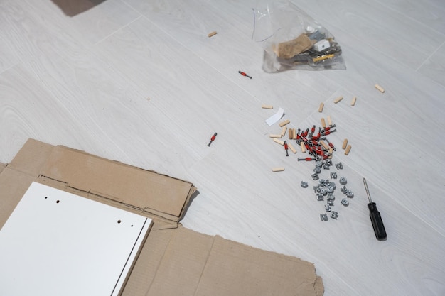 Parts of unfinished furniture, metal screws and tools lying on the floor with instruction manual for furniture assembly in the background. Moving to new home.