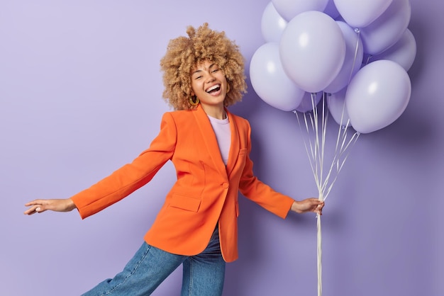 Party is on Cheerful woman dressed in fashionable orange jacket and jeans holds bunch of inflated balloons has joyful expression festive mood isolated over purple background Special occasion