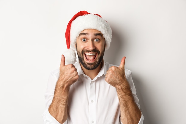 Party, winter holidays and celebration concept. Man enjoying christmas, wearing santa hat and showing thumb up with excited face, white background