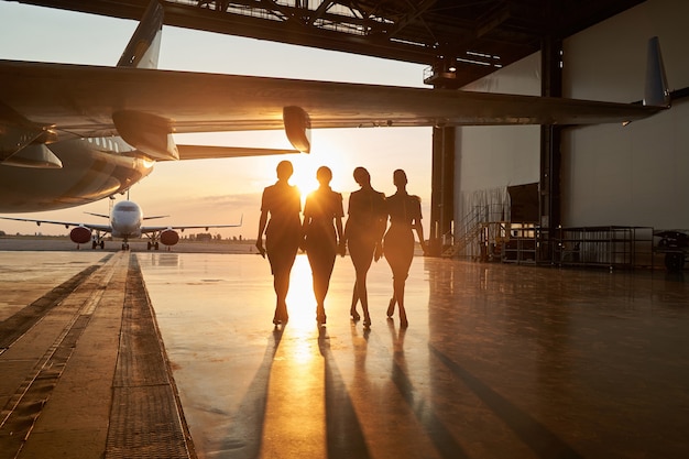 Passenger aircraft in the hangar and four slim female flight attendants walking near it. Big plane outdoors on the background