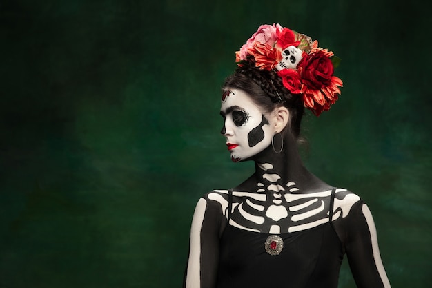 Photo passioned. young girl like santa muerte saint death or sugar skull with bright make-up. portrait isolated on dark green studio background with copyspace. celebrating halloween or day of the dead.