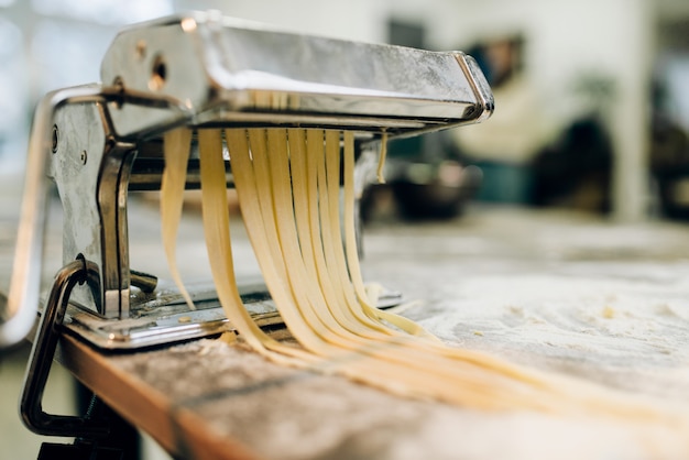 Pasta machine with dough on wooden kitchen table sprinkled with flour closeup. Traditional italian cuisine