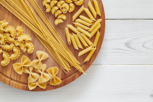 Pasta mix of raw farfalle spaghetti pipe rigatoni maccheroni penne rigate cresta di gallo on round wooden stand on white board background top view space to copy text