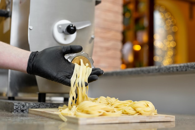 Pasta noodles come out of a professional machine cook opens it and twists it on a wooden board