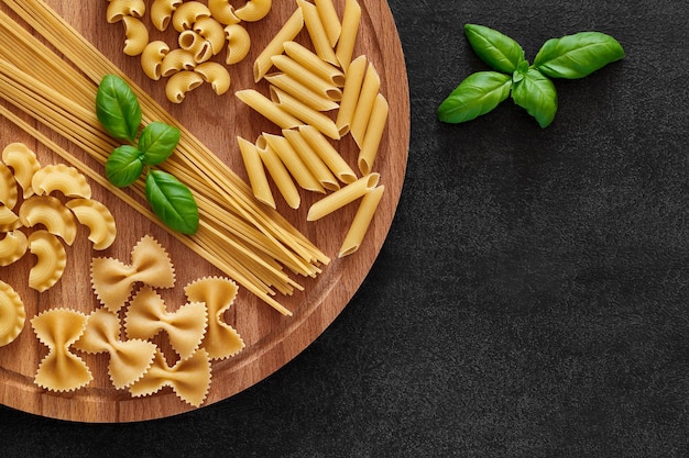 Pasta set of raw farfalle spaghetti pipe maccheroni penne cresta basil leaf on round wooden stand on dark background top view space to copy text