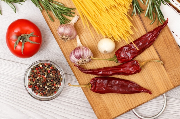 Pasta or spaghetti with ingredients for cooking meat or fish on a wooden cutting board. Garlic, allspice peppers in glass bowl, chili pepper, tomato, onion and rosemary. Top view.