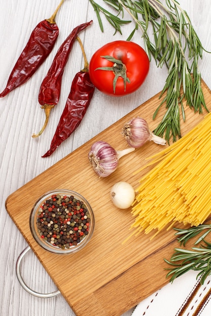 Pasta or spaghetti with ingredients for cooking meat or fish on a wooden cutting board. Garlic, allspice peppers in glass bowl, chili pepper, tomato, onion and rosemary. Top view.