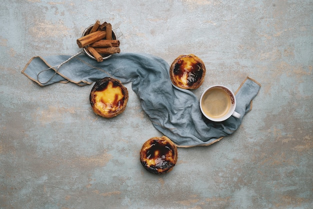 Pastel de nata. Traditional Portuguese dessert, egg tarts over rustic background with cinnamon sticks in the strainer and cup of coffee decorated with napkin. Top view